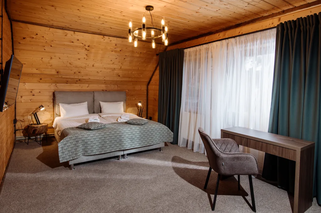 Cozy wooden bedroom with a double bed, bedside lamps, a round chandelier, and a window with sheer curtains.
